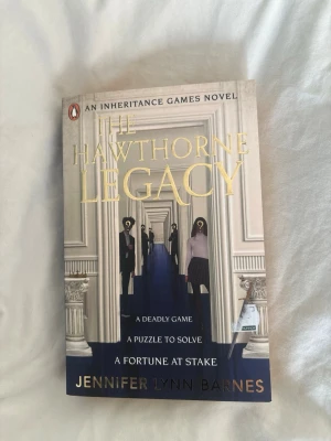 The Hawthorne Legacy pocketbok - Säljer boken 'The Hawthorne Legacy' av Jennifer Lynn Barnes. Omslaget är beige med guldtext och motiv av en korridor med silhuetter. Boken är på engelska och tillhör serien 'An Inheritance Games Novel'. Perfekt för dig som gillar mysterier och spänning.