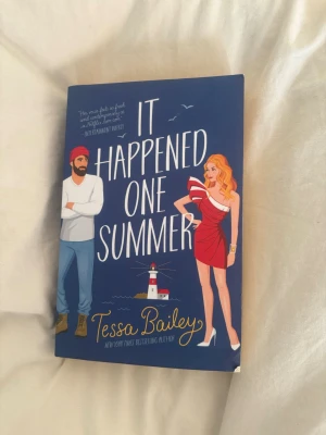 It Happened One Summer bok - En romantisk komedibok på engelska av Tessa Bailey. Omslaget är blått med illustrationer av en man i mössa och en kvinna i röd klänning vid en fyr. Perfekt för dig som gillar feelgood och kärlekshistorier.