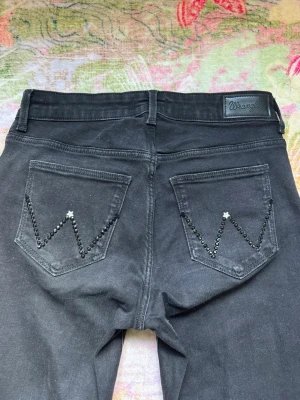 Snygga Svarta Wrangler jeans med pärldetaljer - Vackra svarta bootcut jeans från Wrangler med dekorativa pärlor/glittrande stenar på bakfickorn i W-mönster (Wrangler)😍Klassisk femficksmodell och normal midja! Perfekta för dig som vill ha en unik och mer snyggare plagg som ökar outfiten till topp🩷På framfickorna är det samma dekorativa stenar som matchar ihop hela jeansen. Jättefina jeans som passar till allting, vardagligt och lyxigare events🤍Storlek 27/32 i modellen bootcut, passar XS. Mått cm: midja 33, gren 23, innerben 80. Har stretch❤️