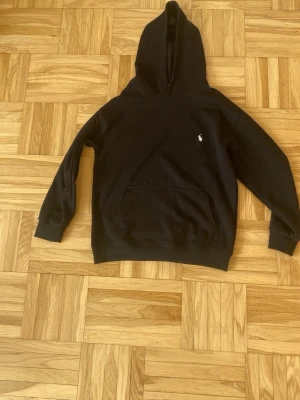 Svart hoodie från Polo Ralph Lauren - Klassisk svart hoodie från Polo Ralph Lauren med huva och magficka. Liten vit logga broderad på bröstet. Tillverkad i mjukt bomullsmaterial som känns skönt mot huden. Perfekt för en avslappnad och stilren look. Det står att den är L fast för mig (183 cm) passar den utmärkt i storlek, fast ärmarna är för små och passar inte min armlängd 