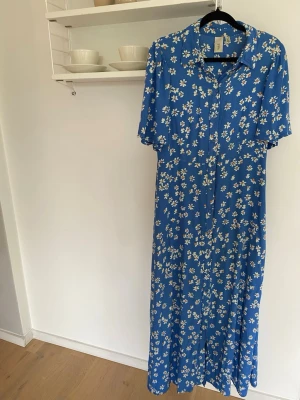 Blå blommig långklänning från YAS - Blå långklänning med vita och gula små blommor, kortärmad och med knappar framtill. Märket YAS.