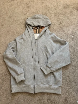 Grå zip hoodie från Burberry - Snygg grå zip hoodie från Burberry med klassiskt rutigt foder i huvan och broderad logga på bröstet. Hoodien har dragsko i huvan, fickor framtill och ribbade muddar. Perfekt för en chill och stilren look.