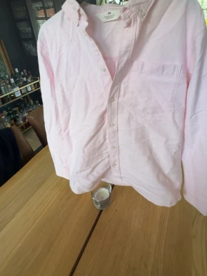 Ljusrosa skjorta från H&M - Säljer en ljusrosa skjorta från H&M med klassisk krage och knäppning framtill. Skjortan har en bröstficka och är tillverkad i mjuk bomull. Perfekt för en avslappnad men ändå stilren look.