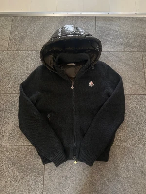 Moncler Backpadded | Cardigan | Hybrid jacka - Snygg svart cardiganjacka med stickade ärmar och quiltad, glansig huva och rygg. Jackan har dragkedja framtill, ribbade muddar och en liten logga på bröstet. Perfekt för kyliga dagar när du vill vara både varm och stilren.