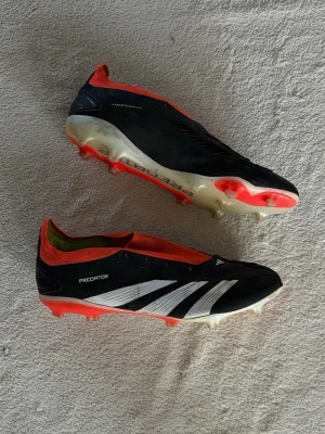 Adidas Predator svart/vit/röd fotbollssko - Säljer ett par Adidas Predator fotbollsskor i svart med vita detaljer och röda inslag. Skorna har låg profil, strumpliknande öppning och mönstrad sula för bra grepp på planen. Tillverkade i syntetmaterial och designade för maximal bollkontroll och komfort.