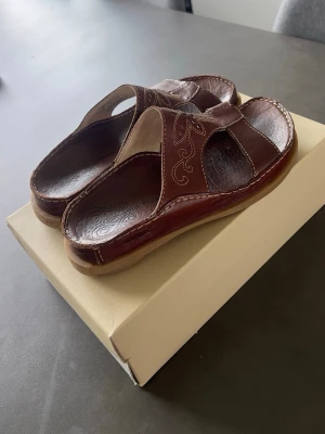 Bruna sandaler med broderi - Säljer ett par bruna sandaler i läderimitation med dekorativa broderier på ovandelen. Sandalerna har öppen tå, breda remmar och en bekväm, räfflad sula i gummi. Perfekta för dig som gillar klassisk och stilren design.