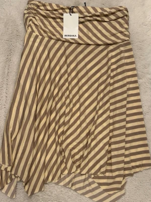 Randig beige och vit kjol från Bershka - Säljer en snygg midilång kjol från Bershka med beige och vita diagonala ränder. Kjolen har en avslappnad passform och är gjord i ett mjukt, stretchigt material som känns skönt mot huden. Perfekt för dig som gillar en chill och trendig stil.