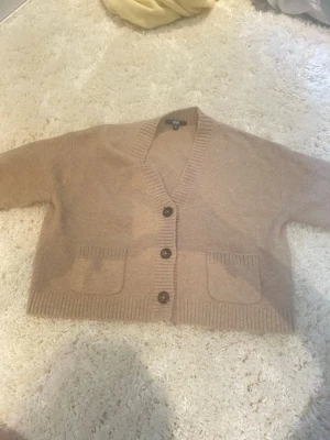 Ljus brun kofta  - Mysig beige kofta med tre stora bruna knappar framtill och två fickor. Koftan har ribbade muddar vid ärmslut, nederkant och runt kragen. Perfekt för lager på lager och enkel att matcha med olika outfits.