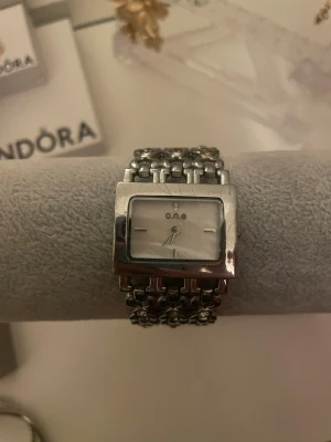 Silverklocka från o.n.e - Stilren klocka från o.n.e med fyrkantig urtavla i vitt och länkarmband i silverfärg. Armbandet har dekorativa detaljer med små stenar som ger en extra touch. Perfekt accessoar för att lyfta din outfit. Klockan fungerar inte men går säkert att laga!!