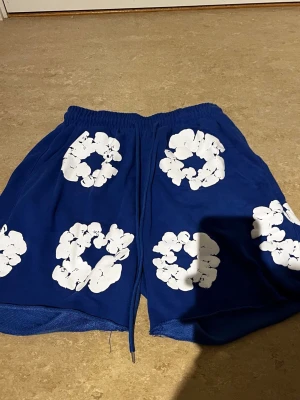Blå shorts med vita blommor från TIm Tears - Blå shorts från Tim Tears med vita blommönster över hela plagget. Shortsen har resårmidja med snörning och är tillverkade i ett mjukt material som passar perfekt för sommaren. Snygg och avslappnad passform.