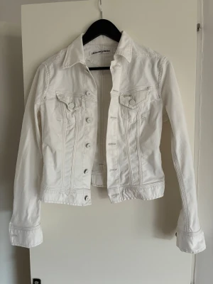 Vit jeansjacka från j. Lindeberg storlek S i nyskick - Snygg vit jeansjackastorleek S från j.Lindeberg med klassisk krage, två bröstfickor med lock och knappar framtill. Figurnära passform och långa ärmar med knapp vid ärmslut. Perfekt att slänga över en t-shirt för en fräsch look.