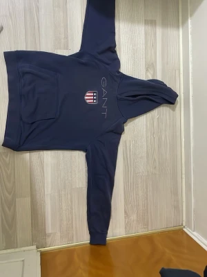 Mörkblå hoodie från GANT - Snygg mörkblå hoodie från GANT med broderad logga och amerikanskinspirerat sköldmärke på bröstet. Klassisk känguruficka framtill och huva med dragsko. Tillverkad i mjukt bomullsmaterial som känns skönt mot huden. Perfekt för en avslappnad och stilren look.