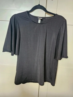 Topp H&M XS - Svart glansig topp med vida ärmar H&M XS  Säljer en svart glansig topp från H&M i storlek XS. Toppen har vida, korta ärmar och en rak passform. Materialet är tunt och skimrande, vilket ger en snygg look. Perfekt för dig som gillar stilrena plagg med det lilla extra.  Kan även hämtas I Partille  E1