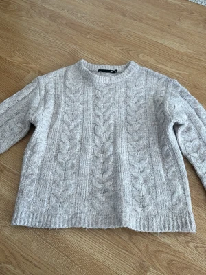 Ljusgrå kabelstickad tröja - Mysig ljusgrå kabelstickad tröja med rund hals och långa ärmar. Tröjan har ett klassiskt flätmönster framtill och på ärmarna, samt ribbade muddar vid ärmslut och nederkant. Perfekt för kalla dagar och enkel att matcha med olika outfits.
