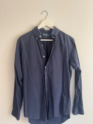 Mörkblå skjorta från Polo Ralph Lauren - Klassisk mörkblå skjorta från Polo Ralph Lauren med button-down krage och broderad logga på bröstet. Skjortan har långa ärmar och vita knappar framtill. Perfekt till jeans eller chinos för en clean look.