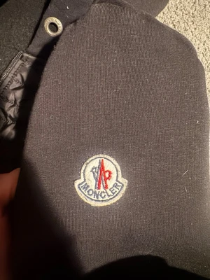 Svart cardigan från Moncler  - Snygg svart hoodie från Moncler med broderad logga på ärmen. Hoodien har dragkedja soppar och quiltad insida i huvan för extra komfort. Perfekt för dig som vill ha en stilren och exklusiv look. Materialet är mjukt och känns lyxigt.
