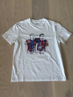 Vit t-shirt med Barcelona-tryck - Vit t-shirt i bomull med tryck på Iniesta och Xavi på bröstet. Motivet har röda och blå ränder samt en gul kaptensbindel på ena spelaren. Klassisk passform och rund hals. Perfekt för dig som gillar fotboll och snygga prints. Vid intresse eller frågor är det bara att höra av sig 