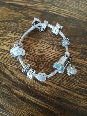 Pandora armband i silver - Säljer ett äkta och snyggt Pandora armband i silver (925) med flera unika berlocker. Armbandet har berlocker i vitt, turkost och lila samt olika former som hjärta, fjäril och blomma. Perfekt för dig som gillar personliga smycken.