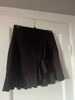 Svart omlottkjol med volang - Säljer en svart kort omlottkjol med volangkant. Kjolen har en snygg och enkel design med en omlott-detalj framtill som ger en extra touch. Perfekt att matcha med en crop top eller stickad tröja för en trendig look.