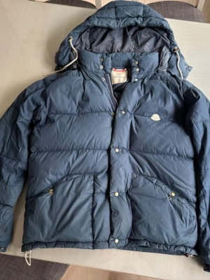 Moncler Mörkblå dunjacka med huva - Snygg mörkblå dunjacka med stor huva och dragsko. Jackan har två stora fickor framtill med knappar och en hög krage med tryckknappar. Klassisk puffer-modell som håller dig varm under kalla dagar. Perfekt för vintern och riktigt chill stil.