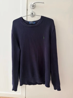 Mörkblå stickad tröja Polo Ralph Lauren - Mörkblå stickad tröja från Polo Ralph Lauren med rund halsringning och diskret grön logga broderad på bröstet. Tröjan har långa ärmar och ribbade muddar vid ärmslut och nederkant. Perfekt för lager på lager och enkel att matcha.