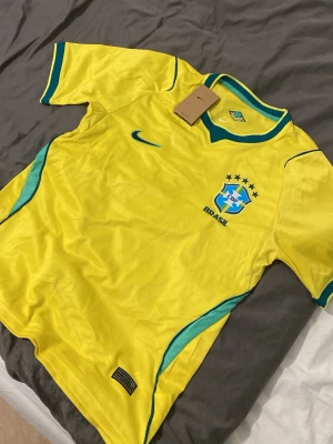 Gul Brasilien fotbollströja Nike - Snygg gul Brasilien fotbollströja från Nike med gröna och blå detaljer. Tröjan har CBF-emblemet broderat på bröstet och klassisk v-ringad krage. Perfekt för fotbollsträning eller att bära när du hejar på landslaget.
