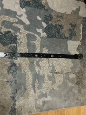 Chrome Hearts belt - Snyggt svart bälte i skinn med silverfärgade nitar och dekorativa detaljer längs hela bältet. Stort ovalt spänne med coolt mönster i silver. Perfekt accessoar för att lyfta din outfit.