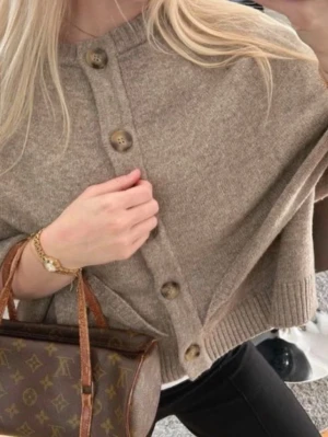 Beige stickad poncho - Snygg beige stickad poncho med oversized passform och stora dekorativa knappar framtill. Tröjan har ribbstickad kant nedtill. Mycket snygg och bra skick!