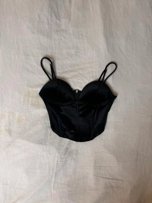 Svart bustier med tunna axelband - Snygg svart bustier med tunna justerbara axelband och vadderade kupor. Modellen har velour aktigt tyg och markerade sömmar framtill för en trendig look. Perfekt att styla under en skjorta eller som den är till höga jeans. Passar XS och S