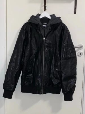 Svart skinnjacka  - Snygg svart skinnjacka med grå huva och dragkedja framtill. Jackan har ribbade muddar, fickor med tryckknappar och en extra ficka med dragkedja på ärmen. Perfekt för dig som vill ha en cool och avslappnad stil. Jag har fler bildr så säg till bara eftersom jag inte kunna ladda upp mer. Mått: längden är ca 73cm, axelbredden är ca 56cm och ärmlängden är ca 70cm långt 💕
