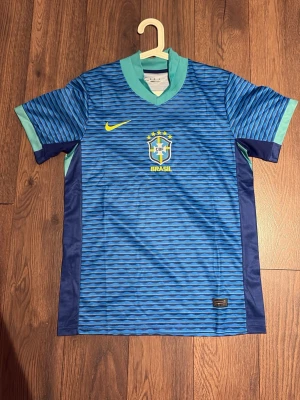 Blå Brasilien fotbollströja Nike - Snygg blå fotbollströja från Nike med Brasilien-emblem på bröstet och gula detaljer. Tröjan har vågigt mönster, ljusblå krage och ärmslut samt mörkblå sidopaneler. Perfekt för dig som älskar fotboll och vill representera Brasilien.