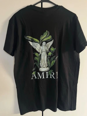 Svart Amiri t-shirt med ängeltryck - Svart t-shirt från Amiri med stort tryck av en vit ängel och gröna blad på ryggen samt mindre tryck på bröstet. Klassisk rund hals och korta ärmar. Materialet ser ut att vara mjuk bomull, perfekt för en clean och stilren look.