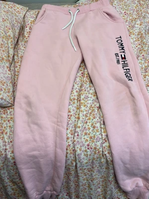 Rosa mjukisbyxor Tommy Hilfiger - Säljer ett par ljusrosa mjukisbyxor från Tommy Hilfiger med tryckt logga och text på ena benet. Byxorna har resår i midjan, vita dragsnören och muddar vid bensluten. Två sidofickor och mjukt material som känns riktigt skönt.