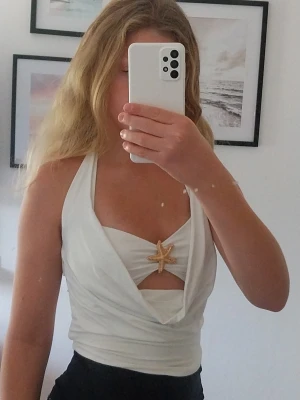 Vit omlottopp med sjöstjärnedetalj - Säljer en vit omlottopp med halterneck och unik sjöstjärnedetalj framtill. Toppen har en öppen design vid bysten och knytband i nacken. Perfekt för dig som vill sticka ut med en somrig och trendig look.