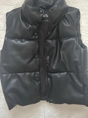 Svart dunväst från ZARA - Snygg svart dunväst från ZARA med hög krage och tryckknappar. Västen har en glansig yta i vad som ser ut som konstläder och quiltad design. Perfekt för lager på lager och ger en cool vibe till din outfit.