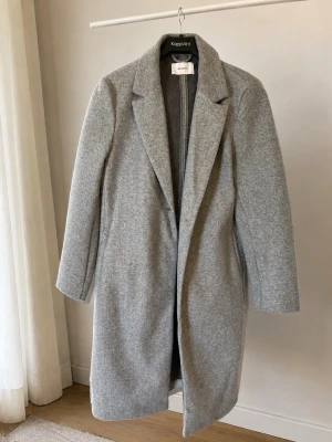 Ljusgrå klassisk ullkappa - Stilren ljusgrå kappa i ullblandning med rak passform och klassisk krage. Kappan har diskreta fickor i sidorna och stängs framtill. Perfekt för dig som gillar minimalistisk och tidlös stil.