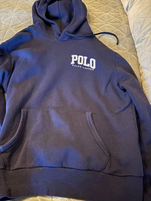Mörkblå hoodie från Polo Ralph Lauren - Snygg mörkblå hoodie från Polo Ralph Lauren med vit logga på bröstet. Klassisk känguruficka framtill och justerbar huva med dragsko. Tillverkad i mjuk bomullsblandning som känns skön mot huden. Perfekt för en avslappnad och stilren look.