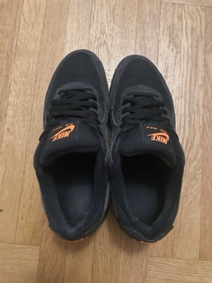 Nike Air Max svarta sneakers - Säljer ett par svarta Nike Air Max 90s  med orange logga på plösen och vit swoosh på sidan. Skorna har klassisk låg profil, svarta skosnören och detaljer i mesh och mocka för en sportig look.