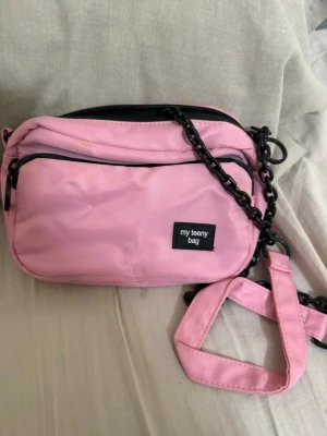 Rosa axelväska från Zara - Snygg rosa axelväska från Zara med svart kedjerem och dragkedja. Väskan har en praktisk ytterficka och en patch med texten 'my teeny bag' på framsidan. Tillverkad i slitstarkt tygmaterial och perfekt för att bära det viktigaste.