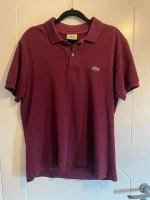 Vinröd pikétröja från Lacoste - Klassisk vinröd pikétröja från Lacoste med korta ärmar och krage. Tröjan har två knappar framtill och den ikoniska gröna krokodilloggan på bröstet. Tillverkad i bomull för en bekväm och luftig känsla.