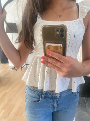 Vit broderad linnetopp med volang - Söt vit linnetopp med breda axelband och volang nedtill. Toppen har broderade detaljer och hålmönster som ger en romantisk känsla. Perfekt till jeans eller kjol för en fräsch sommarlook.