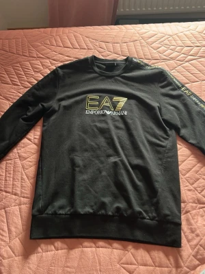 Svart EA7 Emporio Armani sweatshirt - Snygg svart sweatshirt från EA7 Emporio Armani med tryck på bröstet och logga längs ena ärmen. Tröjan har rund halsringning och ribbade muddar vid ärmslut och nederkant. Perfekt för en trendig och sportig look.