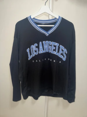 Svart långärmad topp med LA-tryck - Cool svart långärmad topp med blåvitt 'Los Angeles California'-tryck framtill och sportig v-ringad krage med blå och vita ränder. Perfekt för dig som gillar streetstyle och vill ha en avslappnad look.