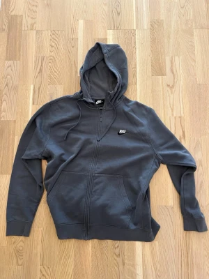 Mörkgrå zip hoodie från Nike - Snygg mörkgrå hoodie från Nike med dragkedja framtill och klassisk huva med dragsko. Broderad Nike-logga på bröstet och två stora fickor framtill. Tillverkad i mjukt bomullsmaterial som är skönt att ha på sig. Perfekt för en avslappnad streetwear-look.