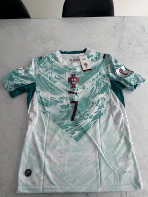 Portugal VM-ställ Ronaldo 7  - Portugal landslagströja från Puma i mintgrönt och vitt med vågigt mönster. Nummer 7 på bröstet, landslagsemblem och Puma-logga. Korta ärmar och DryCell-material som andas. Perfekt för fotbollsträning eller match.
