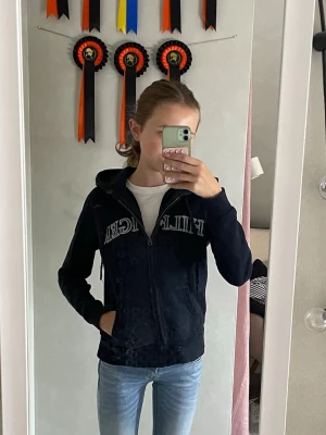 Mörkblå hoodie med dragkedja - Snygg mörkblå hoodie från Tommy Hilfiger med dragkedja framtill och stor logga över bröstet. Hoodien har huva, fickor på sidorna och är tillverkad i ett mjukt material som känns skönt mot huden. Perfekt till jeans eller joggers.