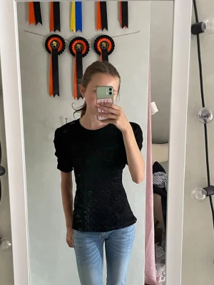 Svart sammetsblus med puffärm - Snygg svart blus i sammet med puffiga korta ärmar och figurnära passform. Perfekt till jeans eller kjol för en trendig look. Materialet ger en lyxig känsla och toppen har en enkel, stilren design.