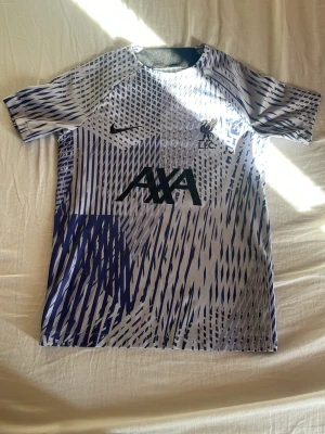 Nike Liverpool FC träningströja mönstrad - Säljer en kortärmad träningströja från Nike för Liverpool FC. Tröjan har ett unikt blått och vitt grafiskt mönster, svart Nike-logga och Liverpool FC-märke på bröstet samt AXA-tryck framtill. Tillverkad i lätt och ventilerande material, perfekt för träning.