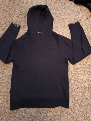Mörkblå hoodie från Tommy Hilfiger - Snygg mörkblå hoodie från Tommy Hilfiger med broderad logga på bröstet. Tröjan har huva, känguruficka och ribbade muddar med diskreta ränder i rött och vitt. Tillverkad i mjukt stickat material som känns skönt mot huden.