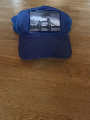 Blå keps med Tower Bridge-tryck - Säljer en blå keps med mesh baktill och justerbar snapback. Framtill finns ett svartvitt tryck av Tower Bridge i London. Kepsen har böjd skärm och är tillverkad i bomull och polyester. Perfekt för dig som gillar streetstyle och coola motiv.